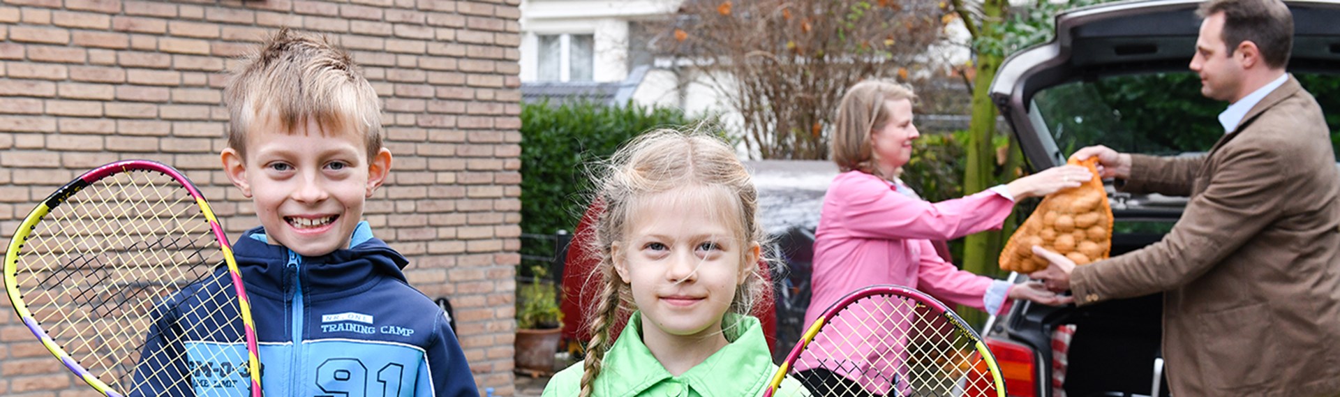 Tochter und Sohn halten Tennisschläger in den Händen und lächeln in die Kamera während im Hintergrund die Eltern das Auto entladen